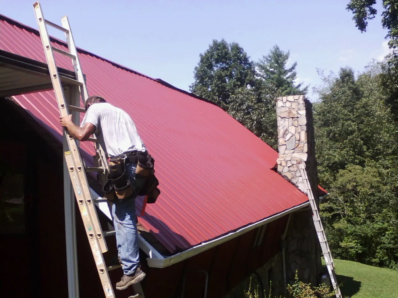 Roofer on a ladder beside a red metal roof and stone chimney — Soffit & Fascia Repair by Roofing Brothers in Plympton